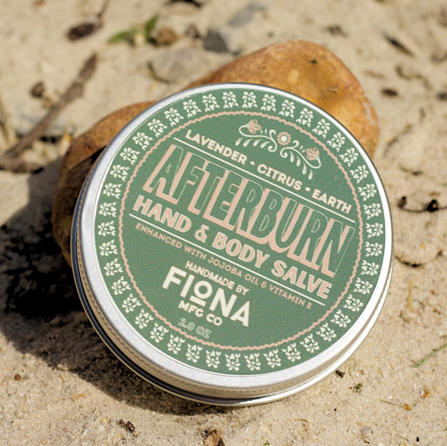 Green and white tin of Afterburn Hand & Body Salve by Fiona on sandy ground